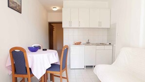 Une pièce compacte comprenant une table à manger, deux chaises, un lit, un coin cuisine avec des meubles blancs et un pare-feu en carreaux.