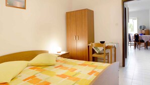Une chambre à coucher avec un lit, une armoire en bois, une petite table avec une chaise, et une vue sur une autre pièce.