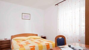 A bedroom with a wooden bed, a nightstand, a chair, a table with a magazine and a glass of water, and a window with sheer curtains. - Holiday apartment with air conditioning (Rabac)