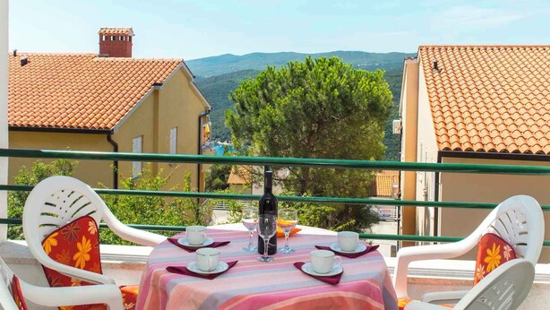 Une terrasse avec une table dressée pour deux personnes, agrémentée d’une bouteille et de verres, entourée de chaises blanches ornées de coussins à motifs floraux.