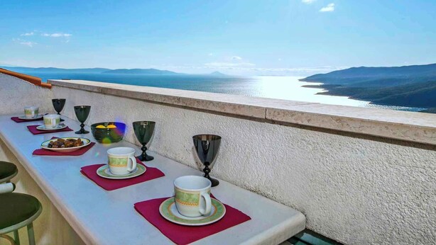 Uma mesa de jantar com pratos, copos e taças numa varanda com vista para a água e colinas ao longe.