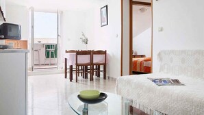 Uma sala de estar com sofá branco, mesa de centro de vidro, área de jantar com cadeiras de madeira e cozinha com televisão e armários.