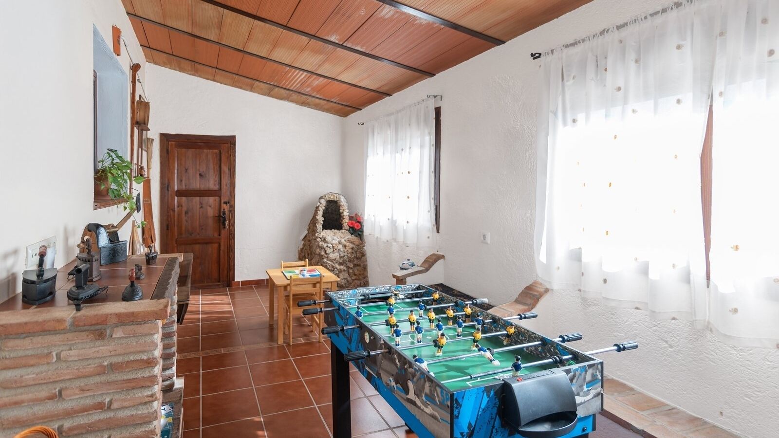 A room with a foosball table, a wooden table with chairs, a stone fireplace, and a door.