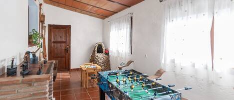 A room with a foosball table, a wooden table with chairs, a stone fireplace, and a door.