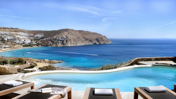 A poolside area with lounge chairs and a view of the sea and a hillside town. - Villa Magnolia  (Mykonos)