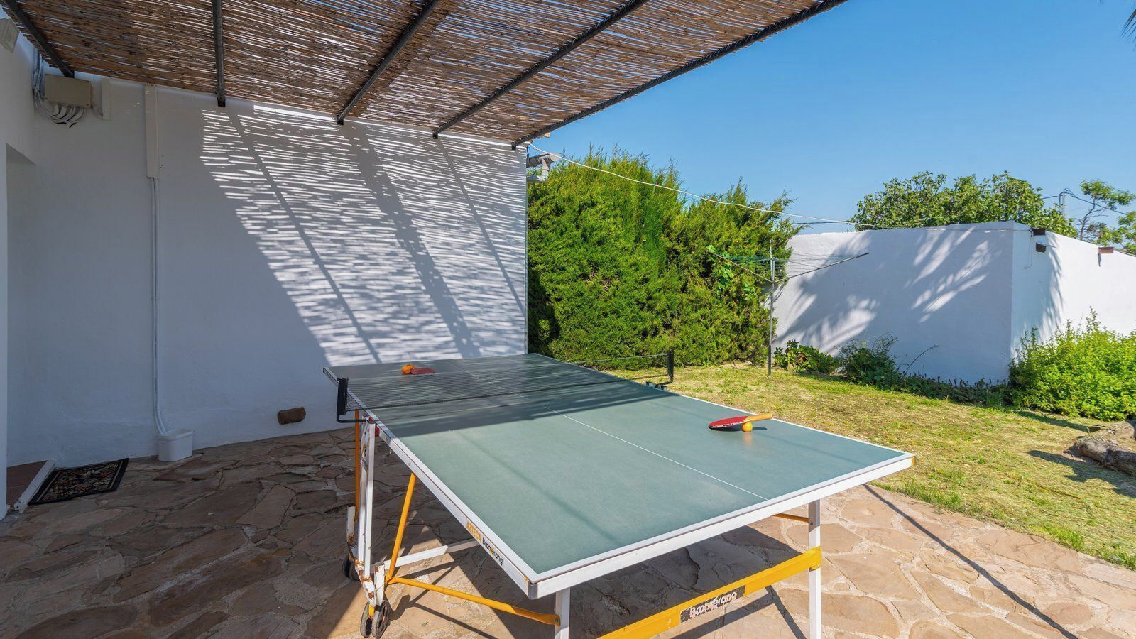 An outdoor ping pong table set up in a garden area with a stone floor, surrounded by greenery and a white wall.