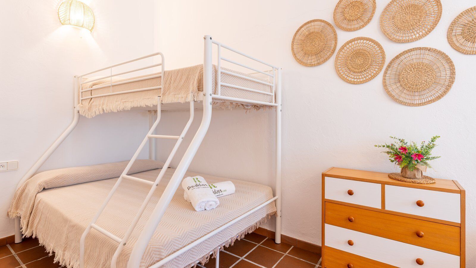 Ein Stockbett mit weißem Metallgestell, eine Holzkommode mit Schubladen und gewebte Wanddekorationen.