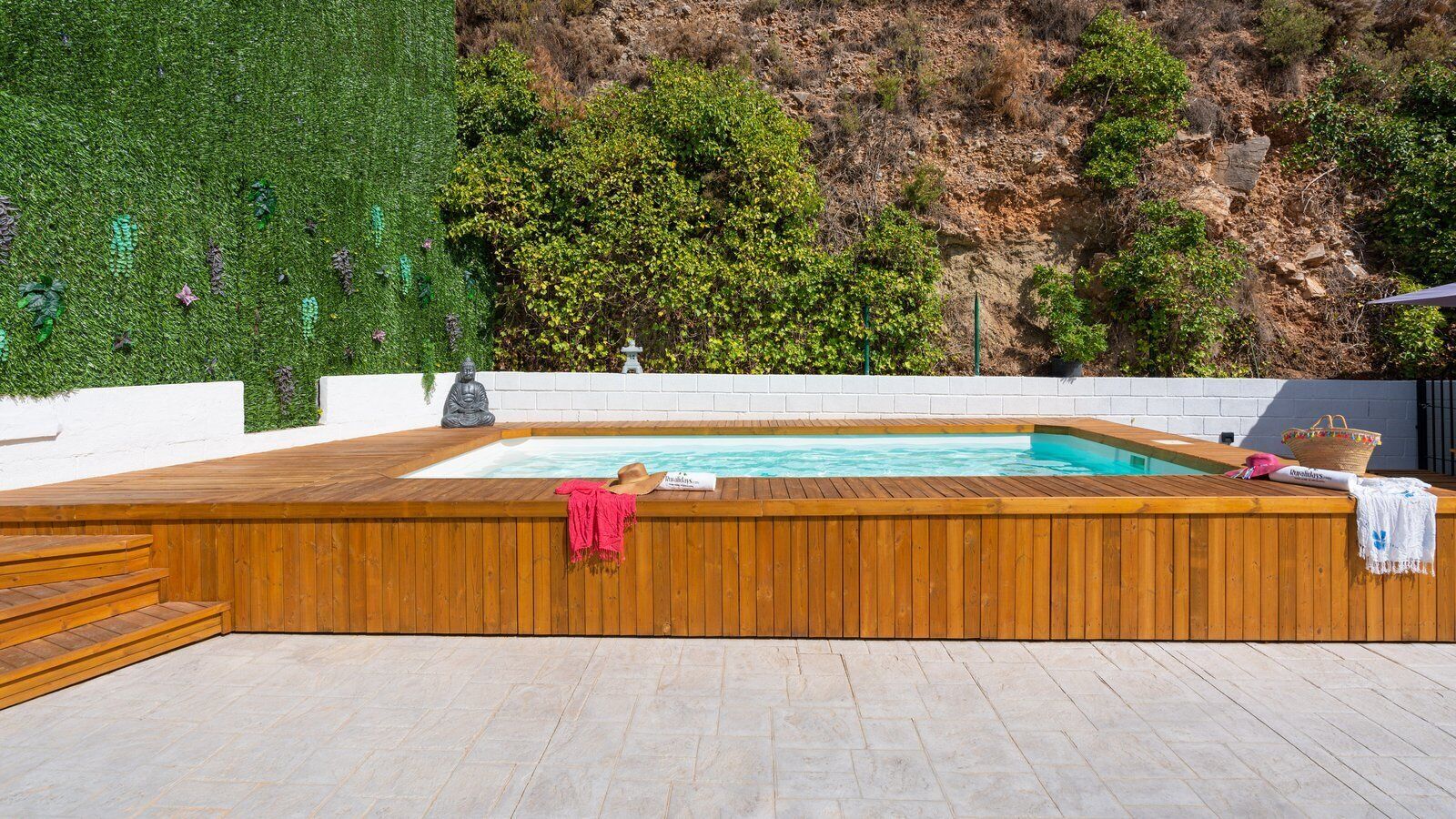 A wooden deck with a hot tub, steps leading up to it, and a white wall with a green hedge behind.