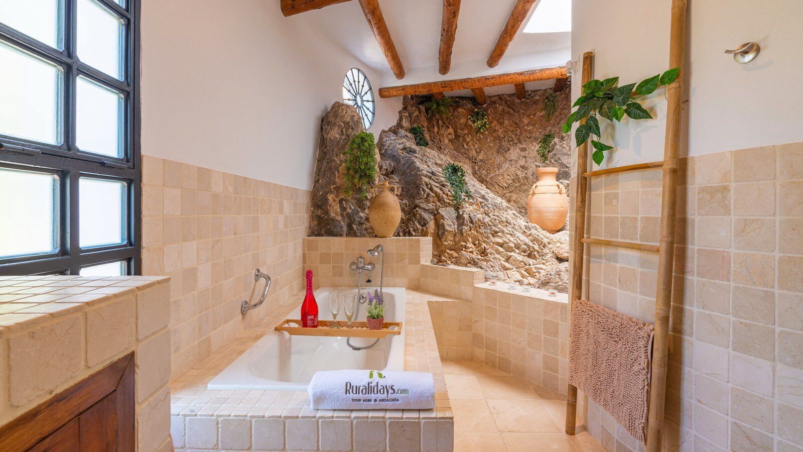 A bathroom with a bathtub, a wooden shelf with bottles, a towel, and a rustic stone wall.
