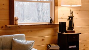 Une pièce chaleureuse avec un canapé, une lampe, une petite table avec des livres et une fenêtre donnant sur des arbres.