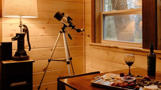 L’intérieur d’un chalet en bois, avec un télescope, une table garnie de nourriture et de vin, et une fenêtre donnant sur la forêt.