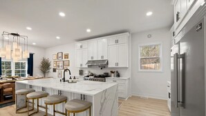 A modern kitchen with white cabinetry, a marble island with bar stools, and a stainless steel refrigerator. - King Beds <> A Sumptuously Lavish Suite Xcape (Alexandria)