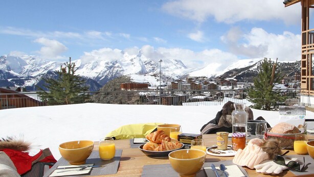 Een ontbijttafel gedekt met croissants, sinaasappelsap en een fles water, met uitzicht op een besneeuwd berglandschap.