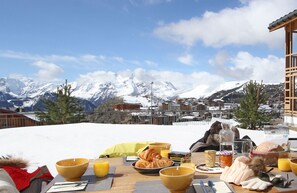 Ein Frühstückstisch mit Croissants, Orangensaft und einer Wasserflasche, mit Blick auf eine verschneite Berglandschaft.
