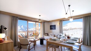 Ein modernes Hotelzimmer mit Essbereich, großen Fenstern mit Bergblick und einem Balkon, im Hintergrund eine Seilbahn.