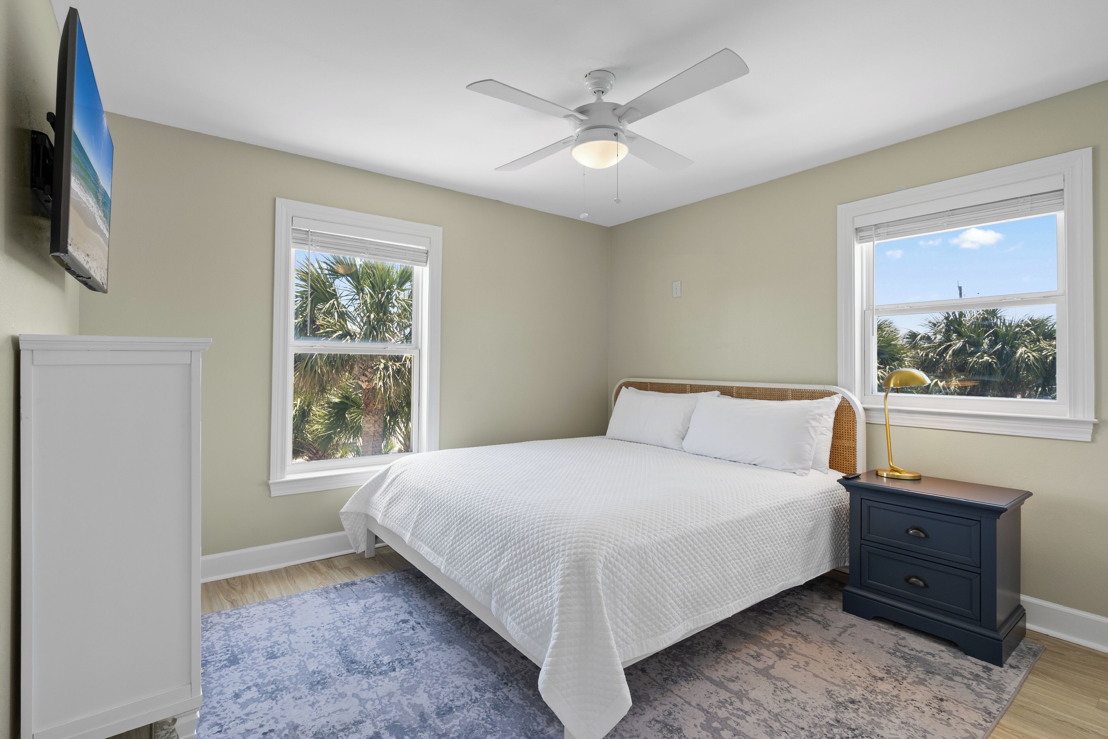 A bedroom with a bed, nightstand, ceiling fan, and two windows.