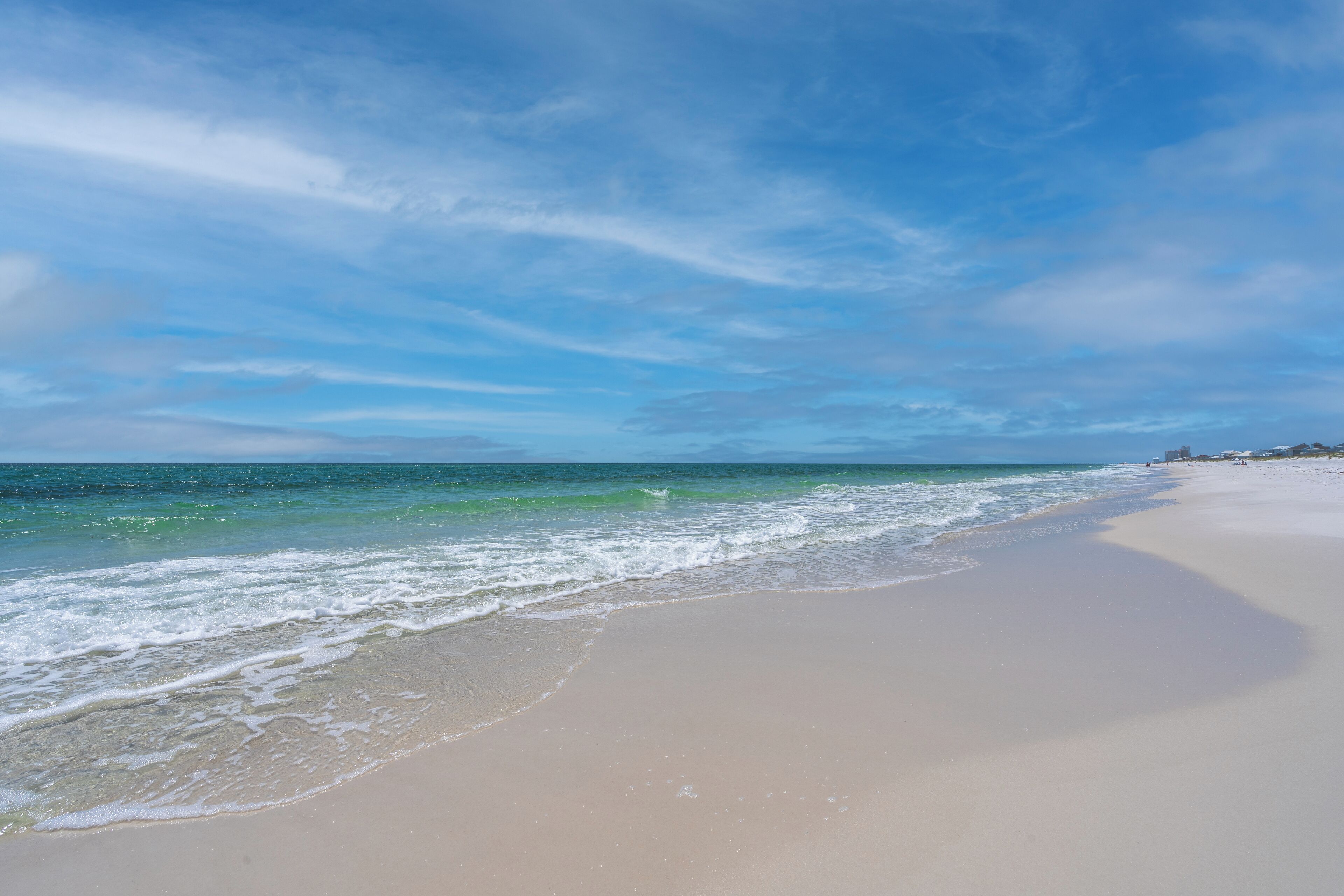A wide sandy beach with gentle waves, clear skies, and distant buildings.