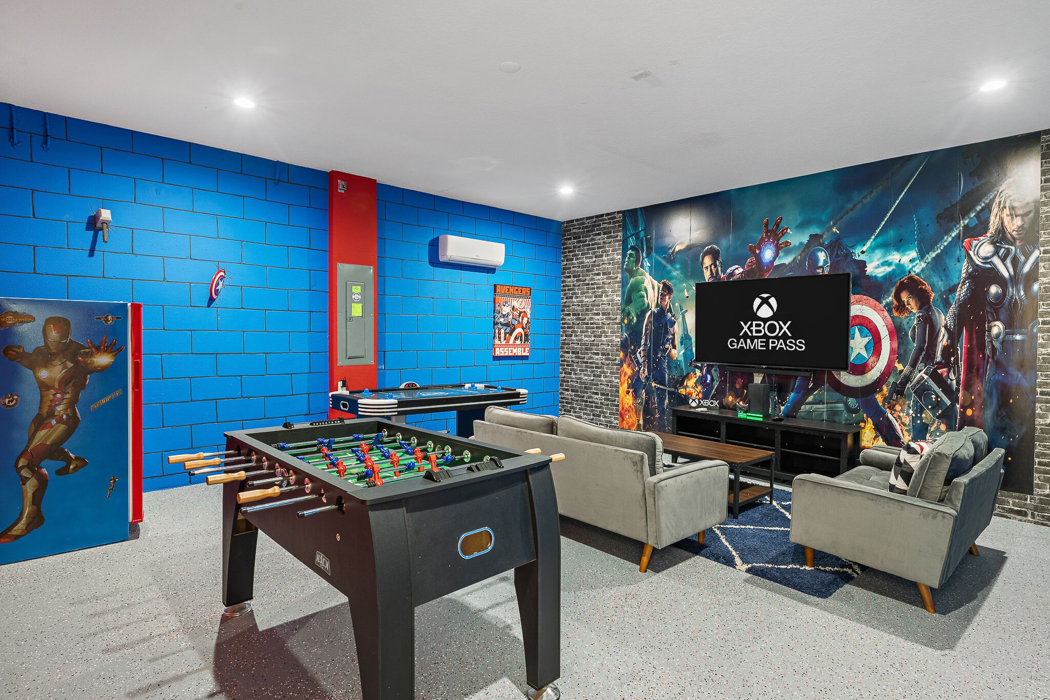 Habitación con mesa de fútbol de mesa, televisor mostrando el logotipo de Xbox Game Pass y un mural con personajes de Vengadores.
