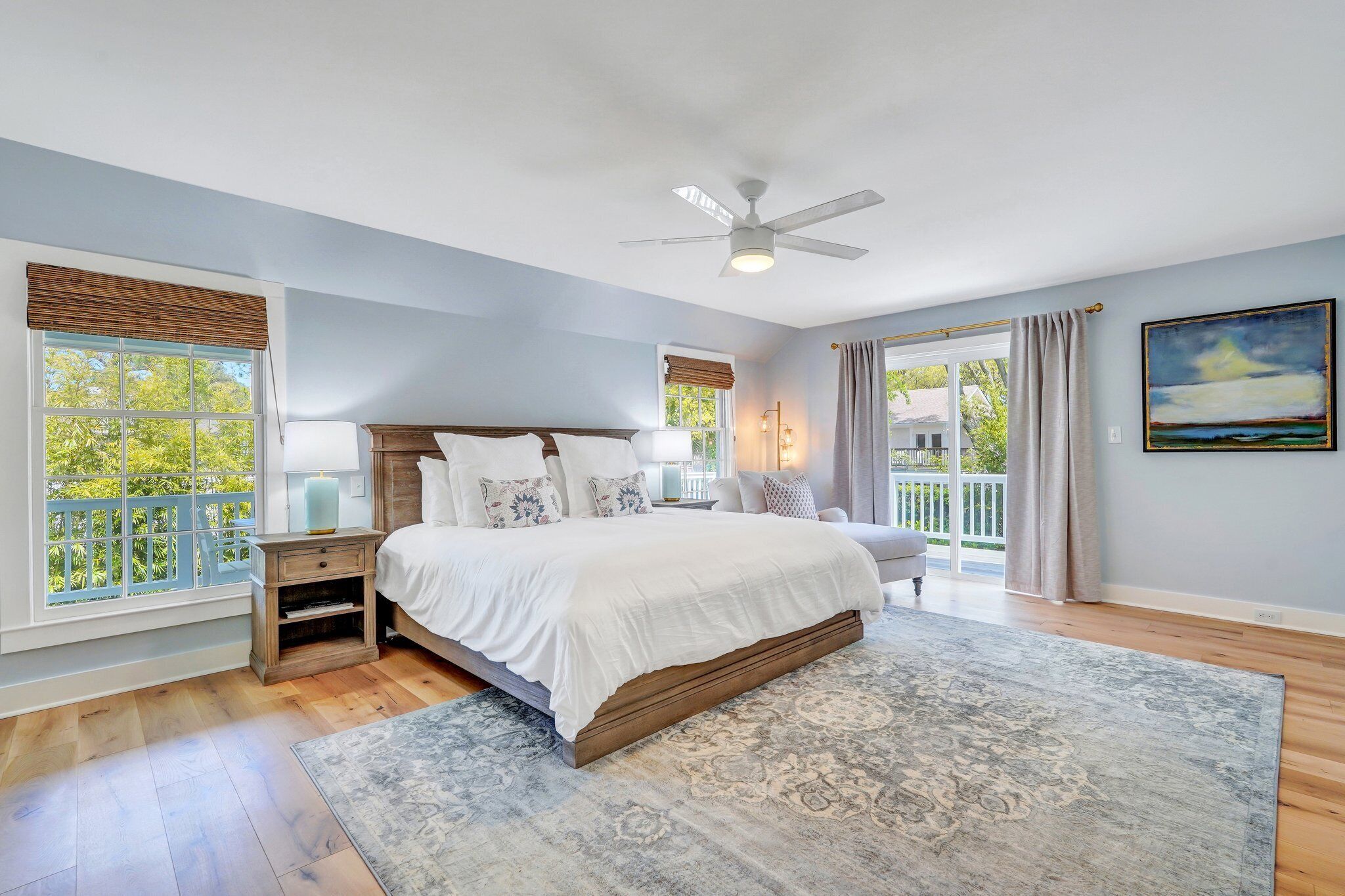 A bedroom with a large bed, a ceiling fan, a nightstand, a lamp, and a view of the outdoors through a window.