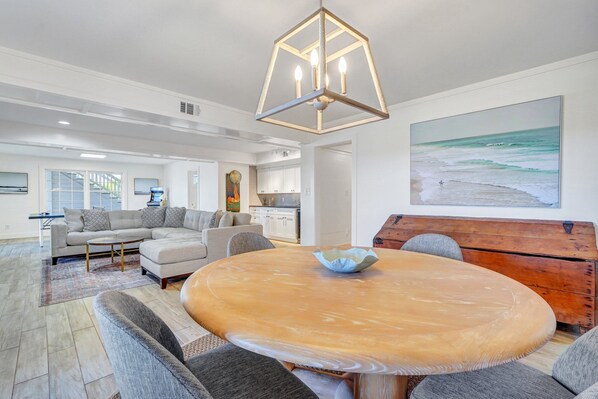A spacious living area with a round wooden table, grey sofas, and a large painting of a beach. - Stay Salty: 6BR East Beach Escape | Pool + Sunset Marsh Views (Saint Simons Island)