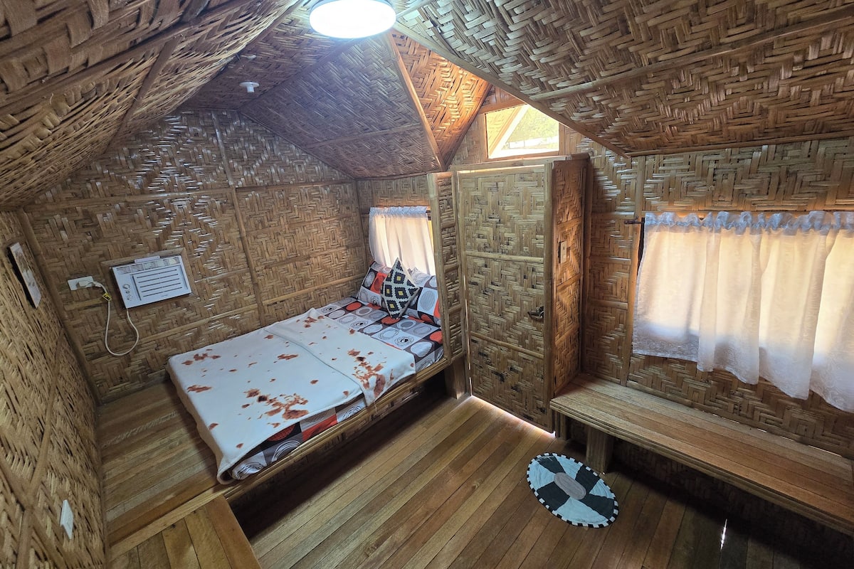 A small, rustic room with a thatched roof, a bed with a patterned bedspread, a bench, and a small table.
