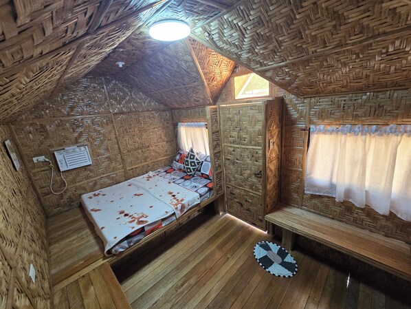 Una habitación pequeña y rústica con techo de paja, una cama con colcha estampada, un banco y una pequeña mesa.