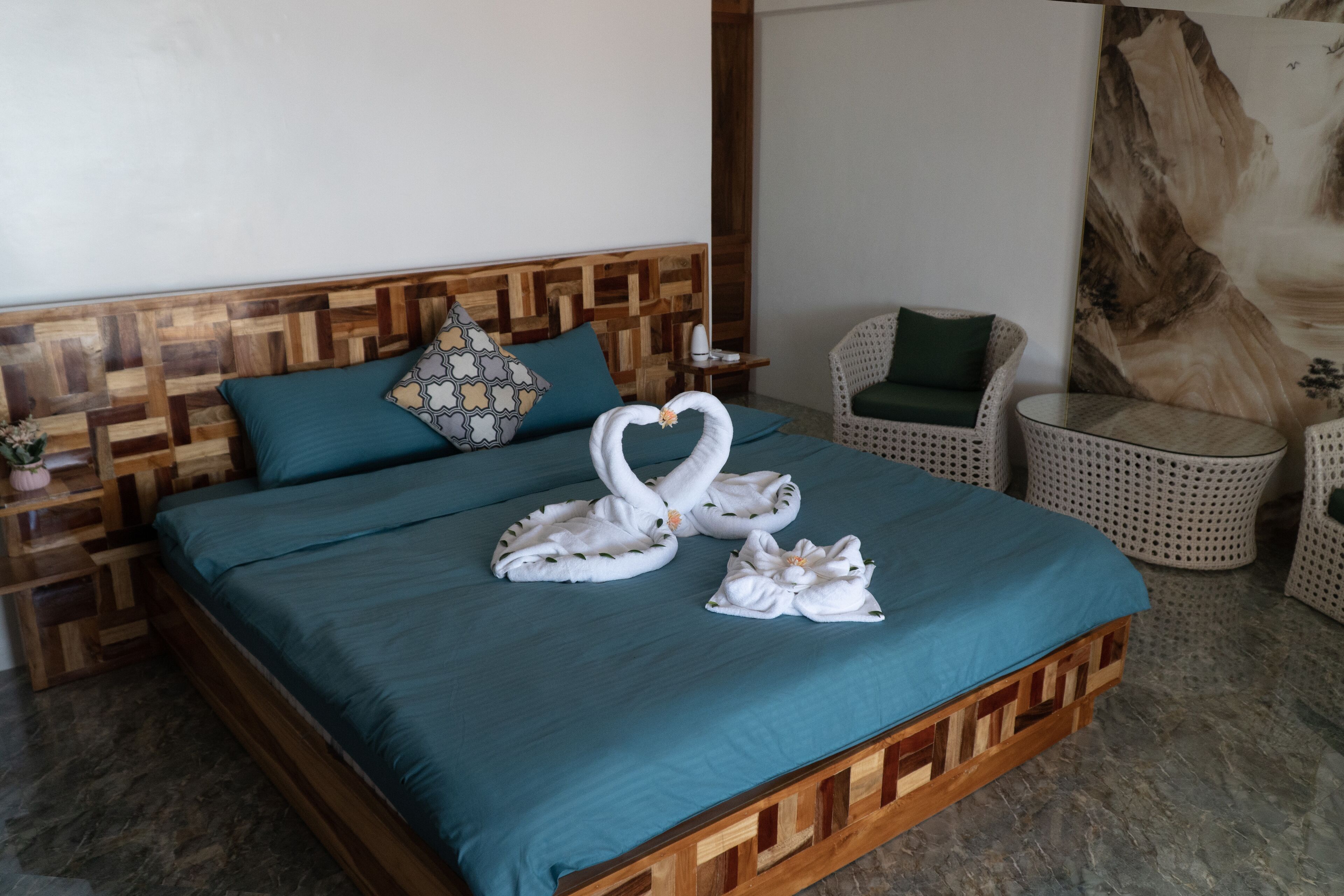 A bed with a blue bedspread, a wooden headboard, and a wall with a wooden panel design. There is a chair with a green cushion and a round table with a white woven seat.