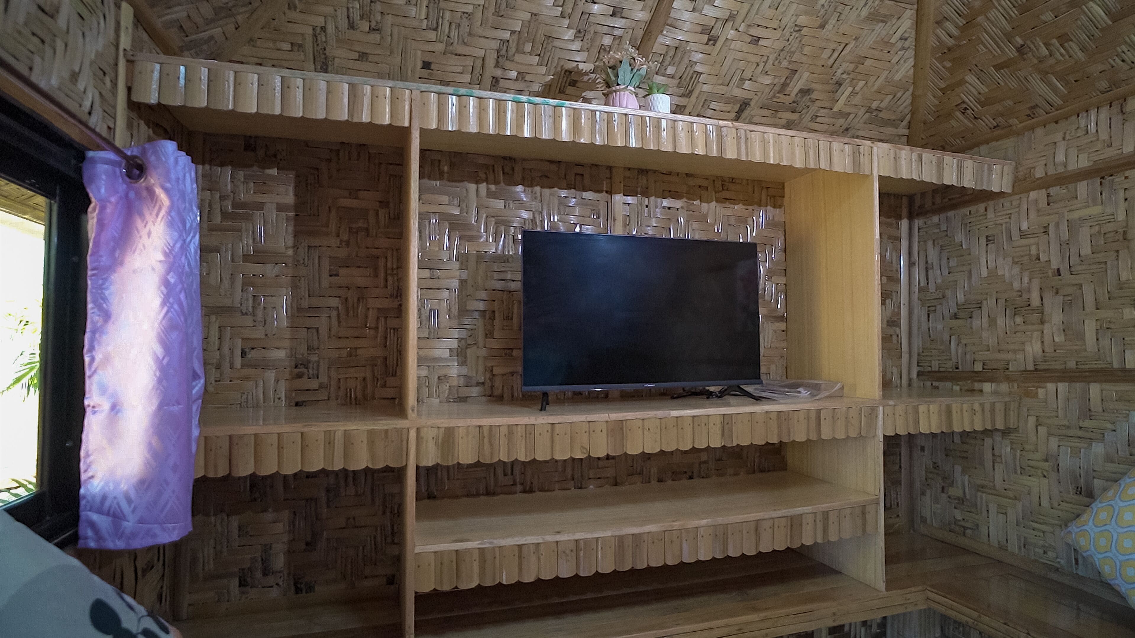 Ein Zimmer mit gewebter Decke, einem Fernseher auf einem Holzregal und einem violetten Vorhang.