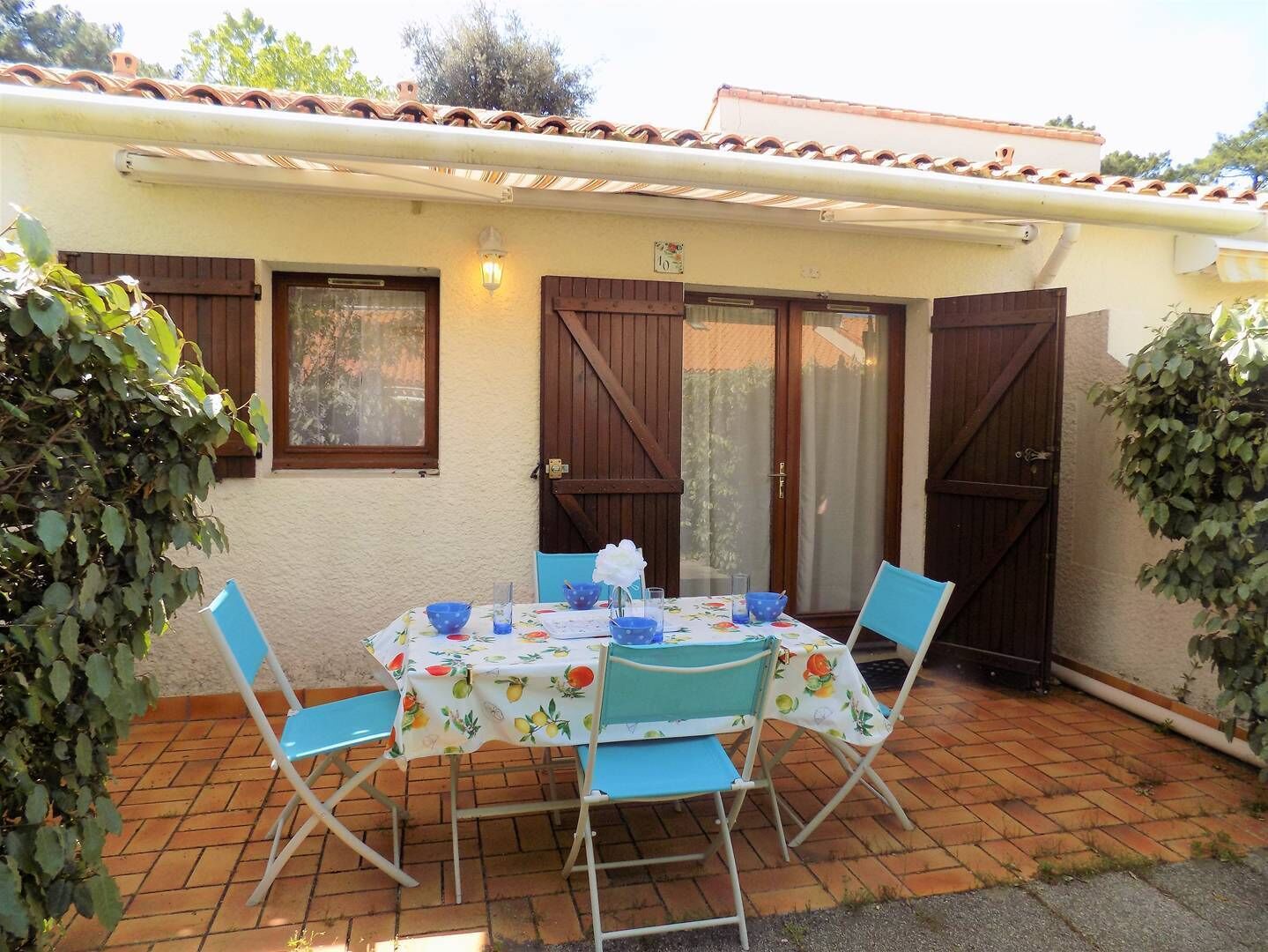 Un patio al aire libre con una mesa preparada para cuatro personas, rodeado de sillas azules, bajo una casa con techo de tejas y contraventanas de madera.