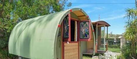 A green covered trailer with a wooden door and windows, a small porch, and a table with chairs outside.