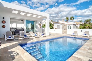 A modern outdoor swimming pool with lounge chairs, a glass door entrance, and a clear blue sky.
