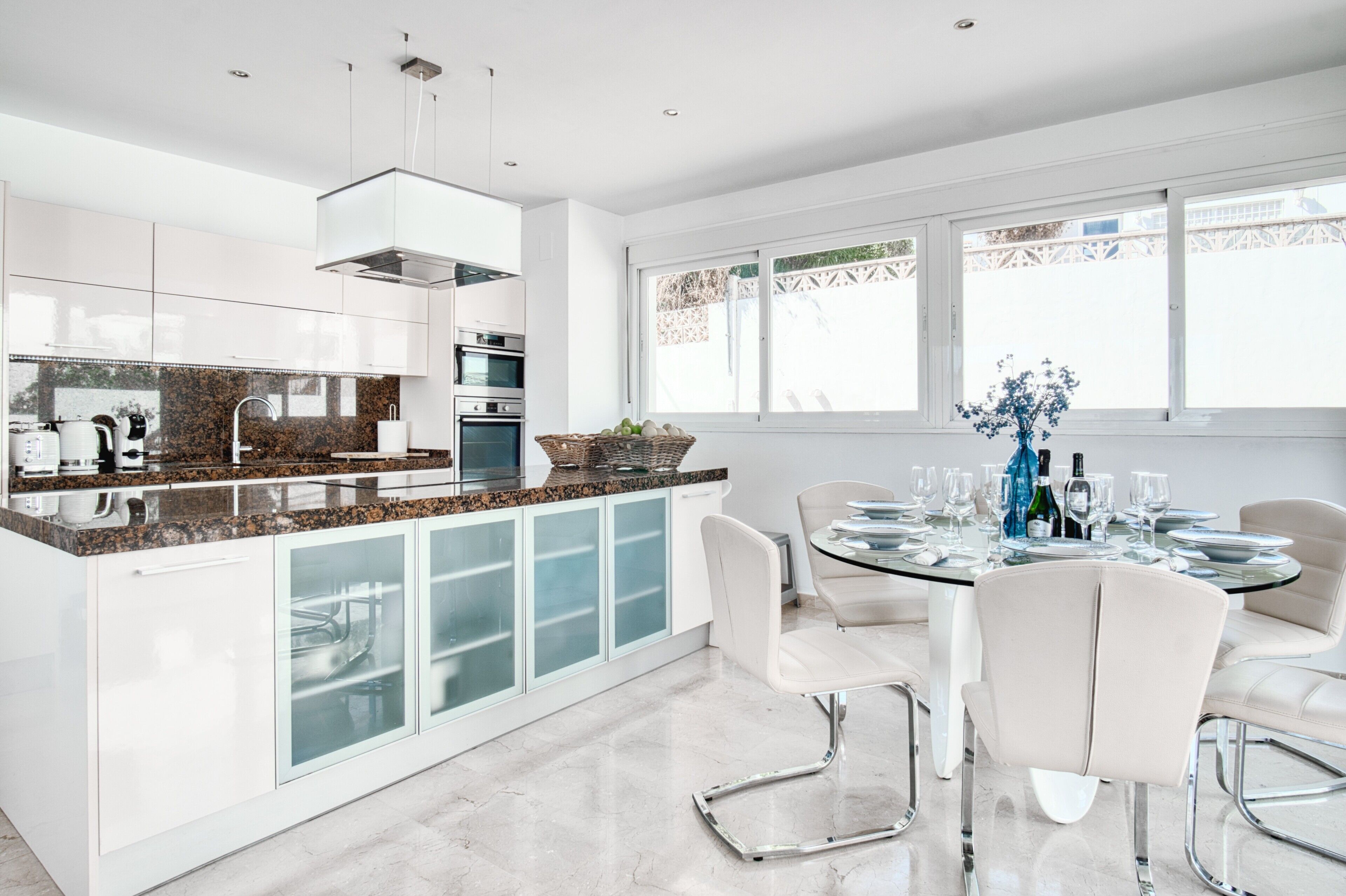 Una cocina moderna con encimeras de granito, armarios blancos y un comedor con mesa de vidrio y sillas blancas.