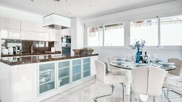 Una cocina moderna con encimeras de granito, armarios blancos y un comedor con mesa de vidrio y sillas blancas.