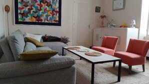 Een woonkamer met een grijze bank, een klein tafeltje met decoratieve borden, een roze fauteuil en een kast met planten.