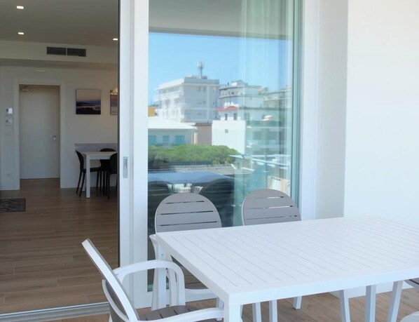 A modern room with a white table and chairs, a sliding glass door leading to a balcony with a city view.