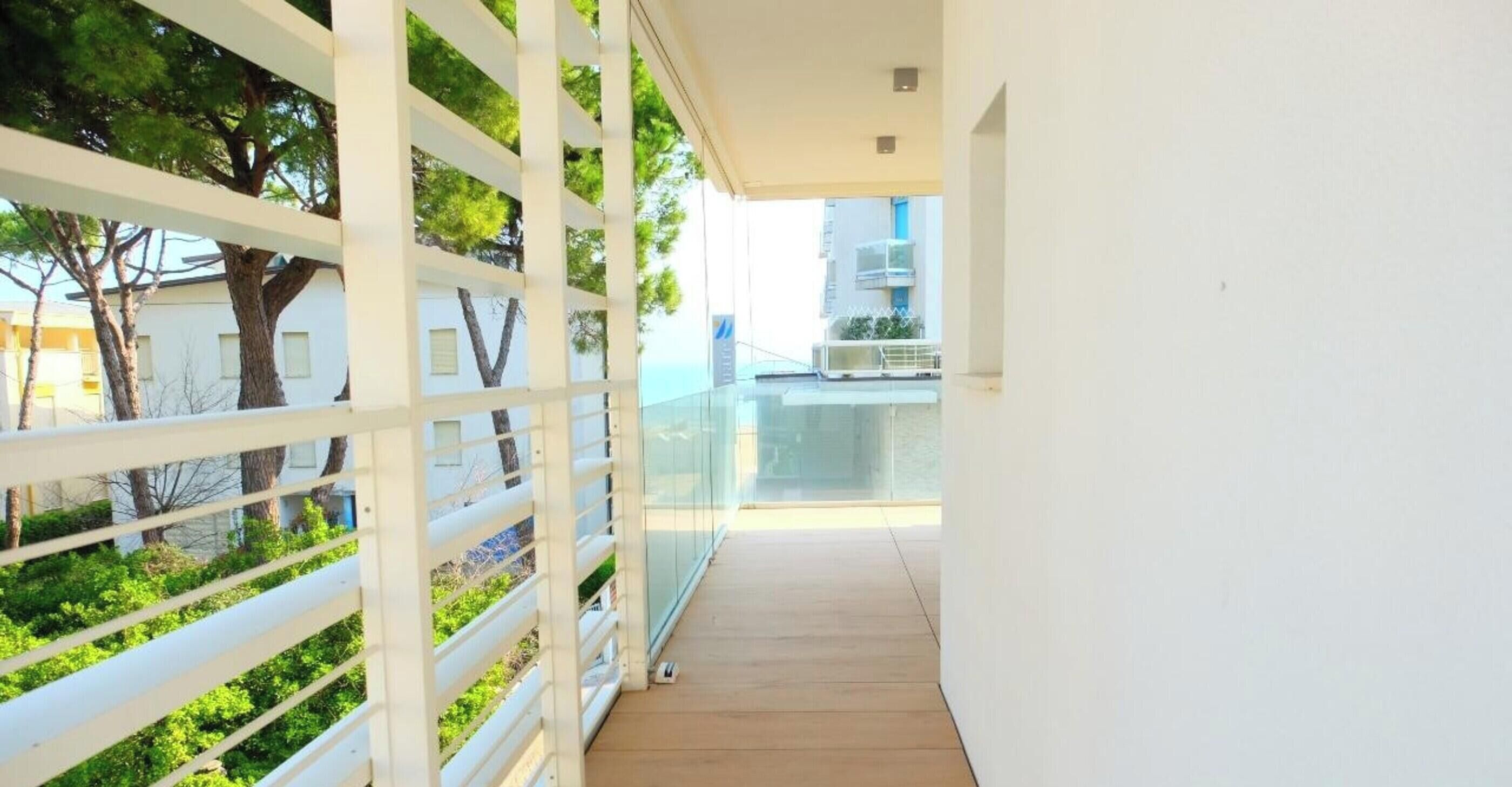 A narrow corridor with a glass railing, white walls, and a view of trees and buildings in the background.