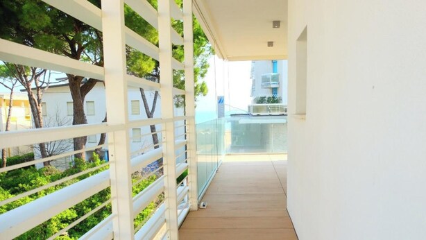 A narrow corridor with a glass railing, white walls, and a view of trees and buildings in the background.