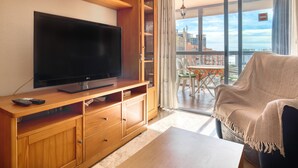 A living room with a flat-screen TV on a wooden cabinet, a sofa, and a balcony with a view of buildings.