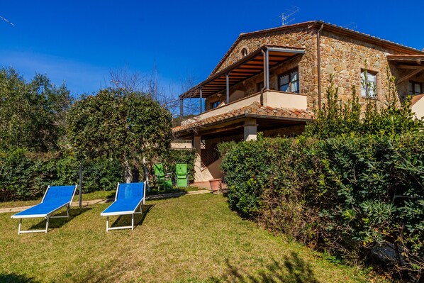 Ein Steinhaus mit Balkon, zwei blauen Liegestühlen und einem gepflegten Garten.