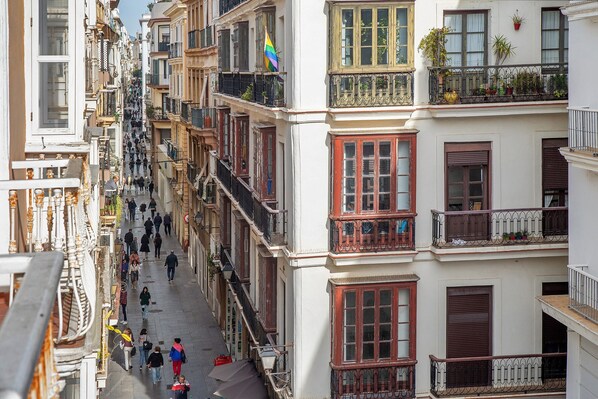 A narrow street lined with multi-story buildings, balconies with plants, and pedestrians.