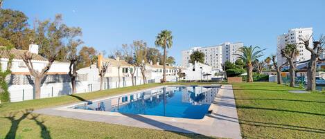 A swimming pool surrounded by grass and trees, with buildings in the background.