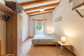 A bedroom with a bed, bedside table, lamp, wooden ceiling beams, and a window with curtains.