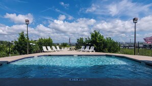 Una piscina con sillas de descanso y una bandera estadounidense, rodeada por una valla y árboles.