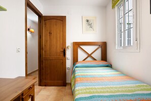 Une chambre avec une porte en bois, un lit recouvert d’une couverture à rayures, une tête de lit en bois, une petite table et une fenêtre avec des stores.