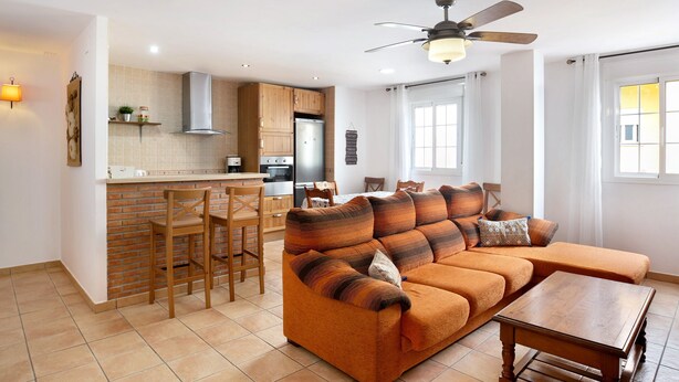 Ein modernes Wohnzimmer mit einer Couch, einem Esstisch und Stühlen. Es gibt eine Küchenzeile mit Schränken und einem Kühlschrank.