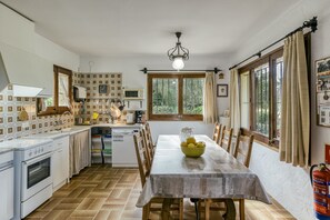 Une cuisine avec une table à manger, des chaises, un saladier de fruits, un extincteur et une fenêtre avec des rideaux.