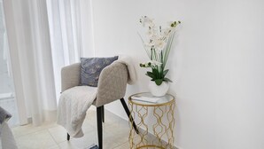 Een stoel met een kussen, een klein rond tafeltje met een gouden draadmotief, en een vaas met witte bloemen.