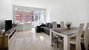 A modern living room with a flat-screen TV, a sofa, a dining table with chairs, and a large window with a view of buildings.