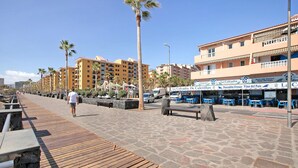 A coastal promenade with a wooden boardwalk, palm trees, and multi-story buildings with balconies.