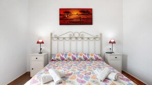A neatly made bed with patterned bedding, two bedside tables with lamps, and a wall-mounted painting of a sunset scene. - Holiday Home 'Casa Ale' with Private Pool, Private Terrace and Wi-Fi (Cádiz)